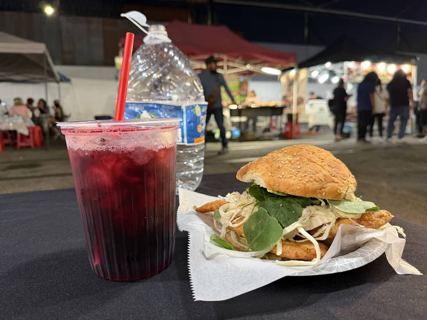 A cemita de milanesa de pollo on a paper plate beside a tall cup of agua de jamaica with a red straw