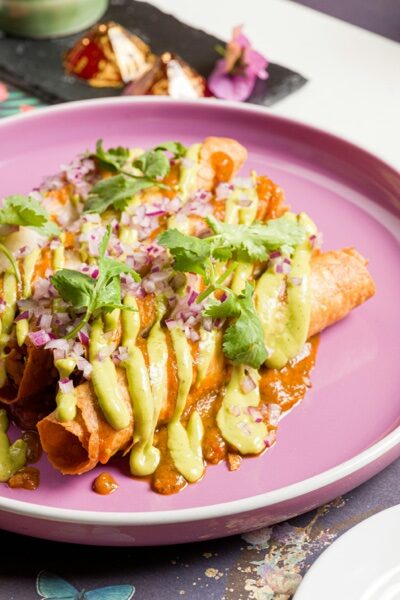 Flautas doradas cubiertas de salsa de aguacate, crema y cebolla morada en plato rosa